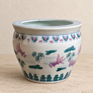 May include: A white ceramic bowl featuring a hand-painted design of pink fish, green pine trees, and clouds. The rim has a blue geometric pattern, while the base is decorated with a green scalloped border. The interior is a smooth, plain white.