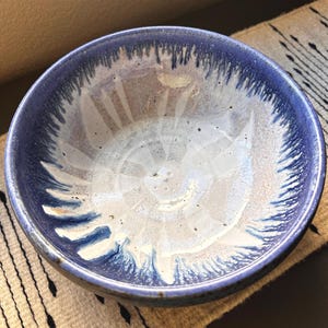 May include: A round ceramic bowl featuring a reactive glaze with a deep blue rim that transitions into a creamy white center. The interior surface displays a distinct drip pattern where the blue glaze bleeds into the lighter base, creating a radial effect.
