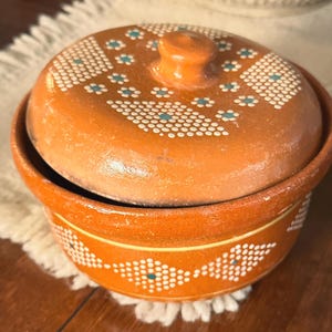 May include: A round terracotta clay pot with a matching lid, featuring a smooth orange finish decorated with white dotted geometric patterns and small teal accents. The lid has a central knob handle, and the pot has a wide, open circular rim.