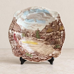 May include: A square ceramic plate with scalloped edges featuring a detailed brown transferware scene of a village with thatched-roof cottages, trees, and a path. The design includes subtle accents of blue in the sky and yellow on the ground.