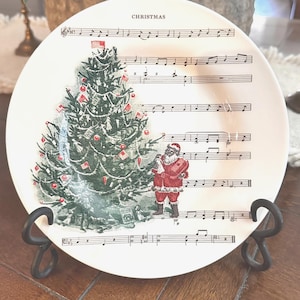 May include: A white ceramic plate featuring a green Christmas tree decorated with red ornaments and small flags, alongside a figure of Santa Claus in a red suit. The background of the plate displays black musical staff lines and the word CHRISTMAS.