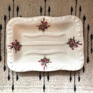 May include: A rectangular ceramic soap dish with a cream-colored base and a scalloped, embossed border. The dish features three horizontal drainage ridges and is decorated with four distinct floral clusters of pink roses and green leaves.