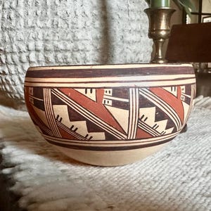 May include: A rounded ceramic bowl featuring a cream-colored base with intricate hand-painted geometric patterns in deep red, black, and white. The design consists of repeating angular shapes, zigzags, and parallel lines encircling the exterior.