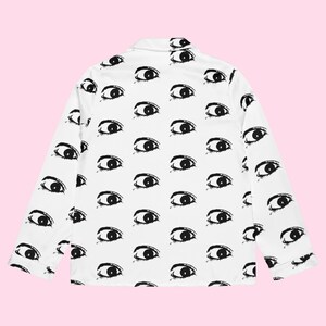 Könnte beinhalten: Weißes Pyjama-Oberteil mit einem sich wiederholenden Muster aus schwarzen Augen. Das langärmlige Hemd hat einen Kragen und eine einzelne Tasche. Das Augenmuster ist gleichmäßig über den Stoff verteilt.