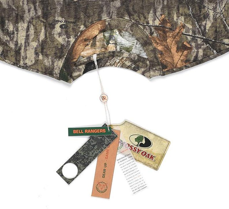 May include: Camouflage pattern garment with tags. The tags include the text "BELL RANGERS", "GEAR UP - GAME", and the Mossy Oak logo. The garment is in shades of brown, green, and tan.
