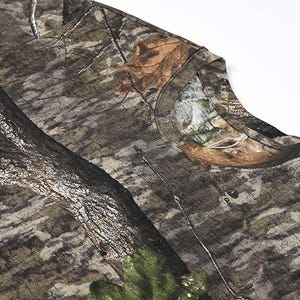 May include: Close-up of a camouflage pattern t-shirt. The fabric features a blend of brown, green, and tan hues, mimicking a forest environment. The design includes tree bark and leaf details, creating a realistic outdoor aesthetic.