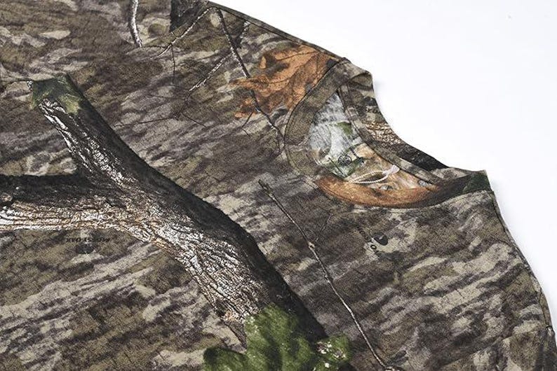 May include: Close-up of a camouflage pattern t-shirt. The fabric features a blend of brown, green, and tan hues, mimicking a forest environment. The design includes tree bark and leaf details, creating a realistic outdoor aesthetic.