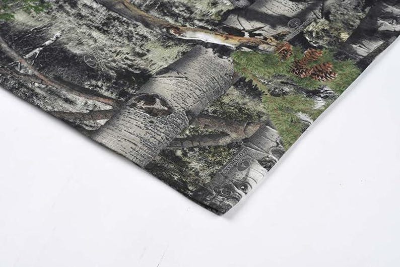 May include: Close-up of a camouflage fabric. The pattern incorporates greens, browns, and greys, resembling a woodland setting. The fabric's texture is evident, with details of tree bark and foliage.