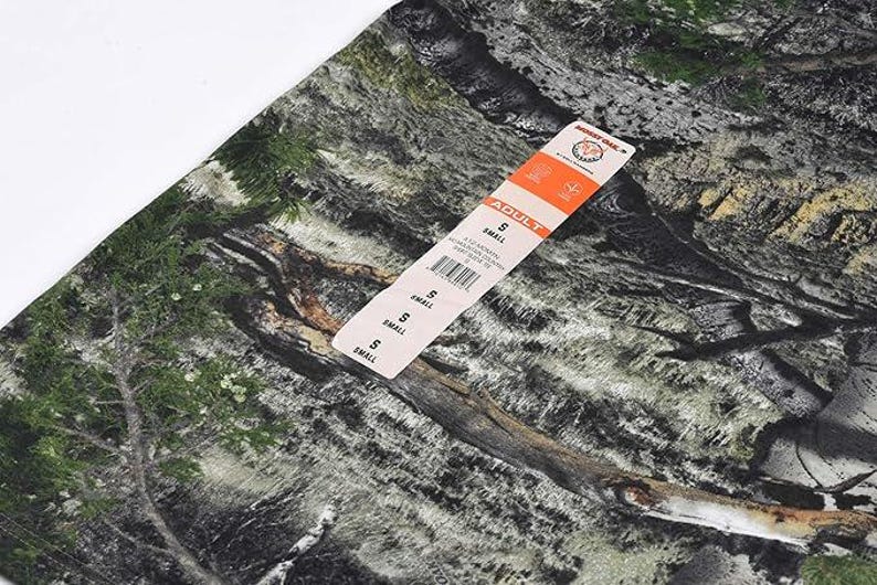 May include: Camouflage fabric in shades of green, brown, and grey, with a tag labelled "ADULT" and "SMALL". The pattern resembles a woodland scene. The tag includes a barcode and a brand logo.