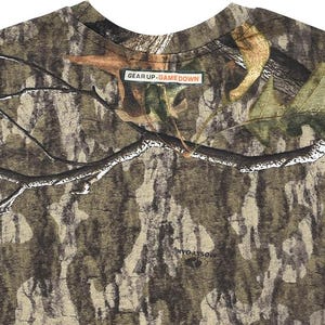 May include: Camouflage pattern t-shirt with a crew neck. The shirt features a brown, green, and tan woodland camouflage design. The text "GEAR UP. GAME DOWN" is printed on a small rectangular label. The brand name "MOSSY OAK" is printed on the shirt.