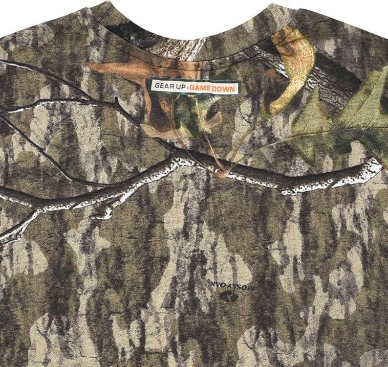 May include: Camouflage pattern t-shirt with a crew neck. The shirt features a brown, green, and tan woodland camouflage design. The text "GEAR UP. GAME DOWN" is printed on a small rectangular label. The brand name "MOSSY OAK" is printed on the shirt.