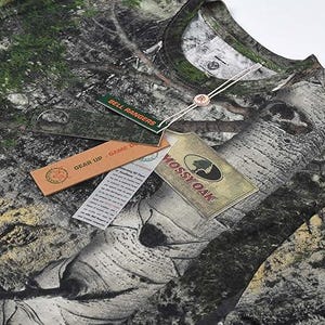May include: A long-sleeved camouflage shirt with a woodland pattern. The shirt has several tags attached, including one that says "Mossy Oak" and another that says "Bell Rangers". The shirt is designed for outdoor activities.