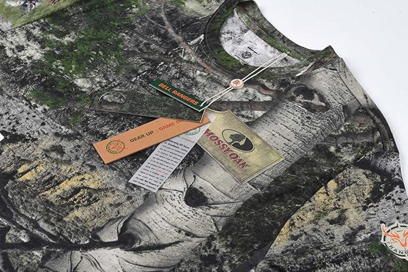 May include: A long-sleeved camouflage shirt with a woodland pattern. The shirt has several tags attached, including one that says "Mossy Oak" and another that says "Bell Rangers". The shirt is designed for outdoor activities.