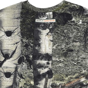 May include: A crew neck t-shirt with a nature-inspired design. The shirt features a detailed print of birch tree bark and foliage in shades of grey, green, and brown. The text "GEAR UP. GAME DOWN" is printed on a rectangular label.
