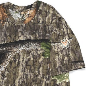 May include: A short-sleeved camouflage t-shirt with a crew neck. The shirt features a brown, green, and tan pattern, with a patch on the sleeve that reads "Rangers" around a deer head logo. The shirt is made of a soft, breathable material.