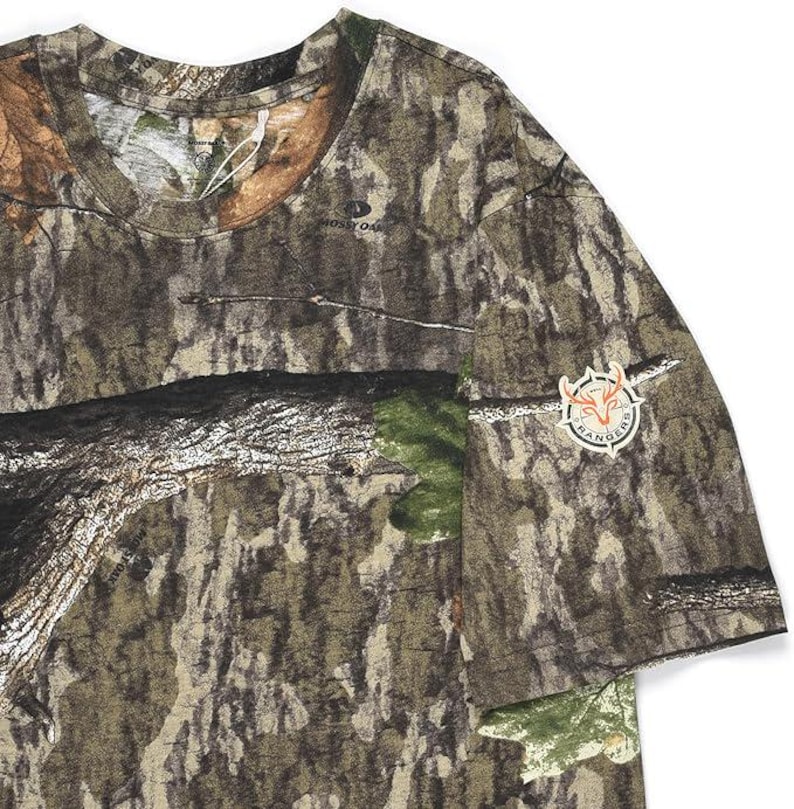 May include: A short-sleeved camouflage t-shirt with a crew neck. The shirt features a brown, green, and tan pattern, with a patch on the sleeve that reads "Rangers" around a deer head logo. The shirt is made of a soft, breathable material.