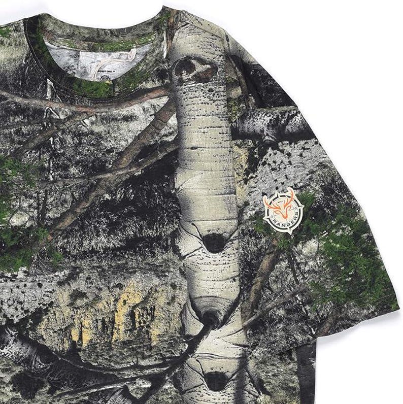 May include: A camouflage-patterned t-shirt with a detailed forest scene print, including trees and foliage. The shirt has a round neckline and short sleeves. A circular patch with the word "Rangers" is on the sleeve.