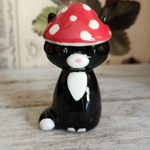 May include: A black cat figurine wearing a red mushroom cap with white spots. The cat has a white bib and a pink nose. The figurine is sitting on a light-colored wooden surface.