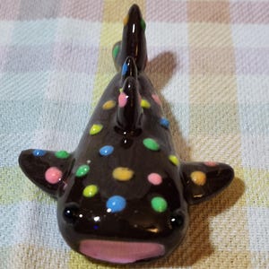 May include: A small, dark brown ceramic whale shark figurine featuring a glossy finish and scattered, raised polka dots in vibrant shades of pink, blue, yellow, and green. The figurine has a wide, flat head with a pink mouth and dorsal fins.