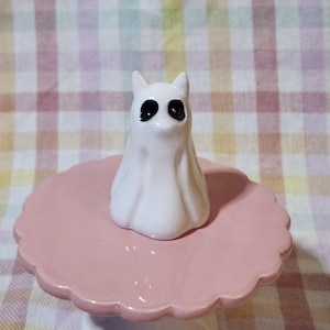 May include: A small white ceramic ghost figurine with two pointed ears and large black oval eyes, positioned on top of a round, scalloped-edge pastel pink pedestal stand with a smooth, glossy finish and a sturdy base.