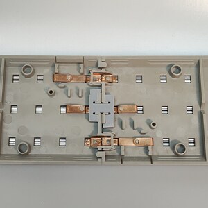 May include: A beige electrical component with copper contacts and a gray plastic center. The component has multiple rectangular slots and circular openings. The copper contacts are arranged in pairs, connected by a gray plastic piece.