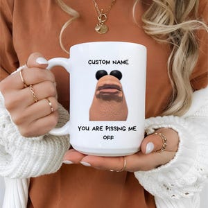 May include: White ceramic mug with a handle, featuring a humorous graphic of a face with sunglasses and the text "CUSTOM NAME" above and "YOU ARE PISSING ME OFF" below. The mug is held by a person.