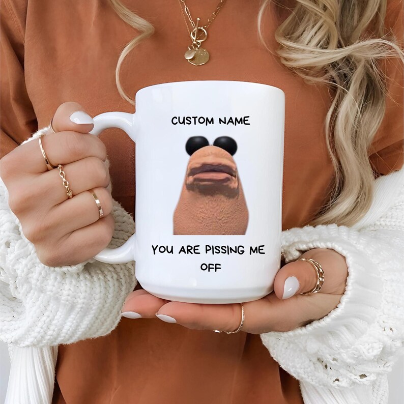 May include: White ceramic mug with a handle, featuring a humorous graphic of a face with sunglasses and the text "CUSTOM NAME" above and "YOU ARE PISSING ME OFF" below. The mug is held by a person.