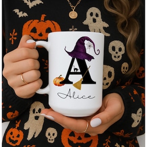 May include: White ceramic mug with a Halloween-themed design. The design includes a purple witch hat, a black cat, a broom, and a pumpkin. The letter "A" is prominently displayed, with the name "Alice" written below.