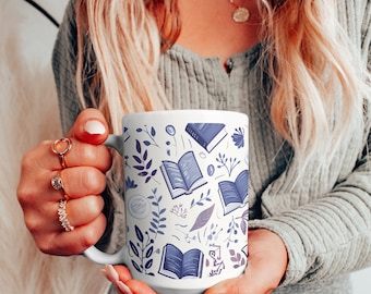 May include: White ceramic mug with a blue and white pattern of open books and floral designs. The mug is being held by a person's hand.