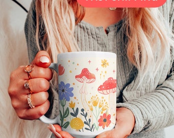 May include: White ceramic mug with a colorful floral and mushroom design. The design features red, yellow, blue, and green flowers and mushrooms. The mug has a handle and is held by a person's hand.