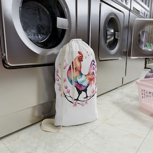 May include: A white laundry bag with a colorful rooster illustration and pink cherry blossoms. The bag is sitting in front of a row of washing machines.