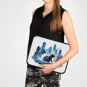 May include: A white laptop sleeve with a watercolor illustration of a city skyline and a Godzilla-like monster emerging from the water. The sleeve has a zipper closure.