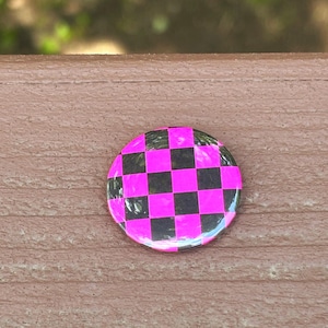 May include: A circular pin-back button featuring a vibrant checkerboard pattern in alternating bright neon pink and solid black squares. The surface of the button has a smooth, glossy finish that reflects light across the geometric grid design.