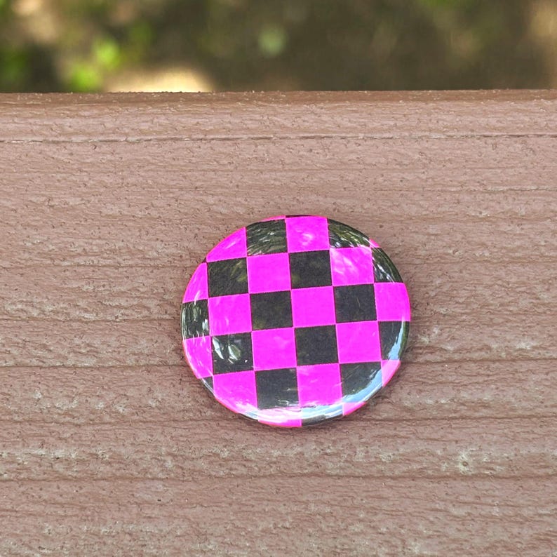 Peut inclure: Un badge rond avec un motif &agrave; damier compos&eacute; de carr&eacute;s altern&eacute;s rose n&eacute;on vif et noir. La surface du badge pr&eacute;sente une finition lisse et brillante qui refl&egrave;te la lumi&egrave;re sur l'ensemble du motif g&eacute;om&eacute;trique en forme de grille.