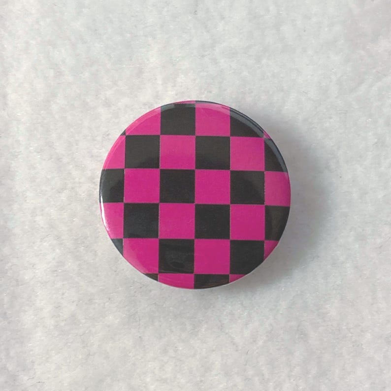 Peut inclure: Un badge rond avec un motif &agrave; damier r&eacute;p&eacute;titif compos&eacute; de carr&eacute;s noirs et de carr&eacute;s rose vif altern&eacute;s. La surface du badge est lisse et brillante, pr&eacute;sentant un design g&eacute;om&eacute;trique &agrave; grille avec un contraste de couleurs marqu&eacute;.