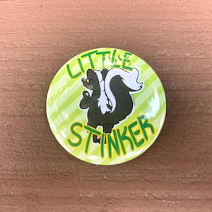 May include: A circular pinback button featuring a light green and white diagonal striped background. In the center is a cartoon illustration of a black and white skunk with the text LITTLE STINKER written in green letters above and below it.