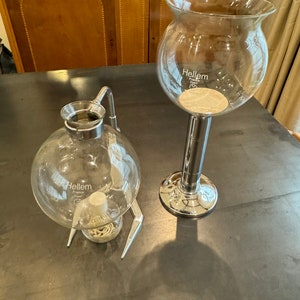 以下が含まれることがあります： クロムのアクセントが付いたガラス製のコーヒーメーカーが2つ。大きなコーヒーメーカーは丸いベースと背の高い円筒形のステムを持っています。小さなコーヒーメーカーは丸いベースと湾曲したステムを持っています。両方のコーヒーメーカーには「Hellem」という文字が印刷されています。