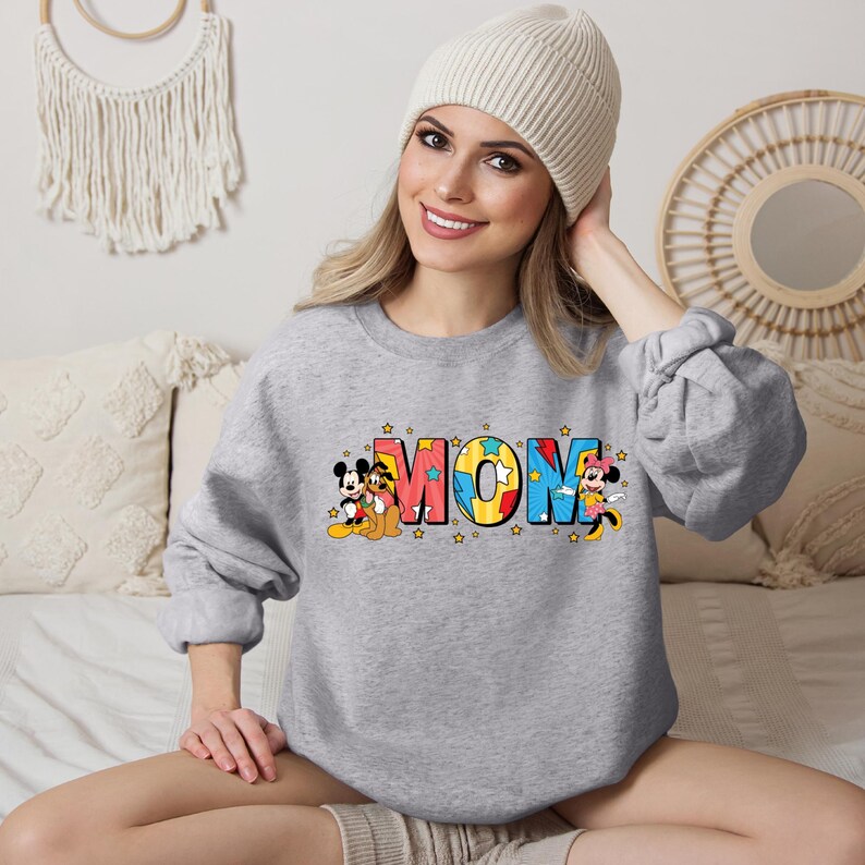 K&ouml;nnte beinhalten: Grauer Pullover mit einem bunten "MOM"-Motiv mit Mickey Mouse, Minnie Mouse und Pluto. Die Buchstaben sind schwarz umrandet und mit verschiedenen Farben gef&uuml;llt, darunter Rot, Blau, Gelb und Gr&uuml;n. Um das Motiv herum sind kleine gelbe Sterne verteilt.