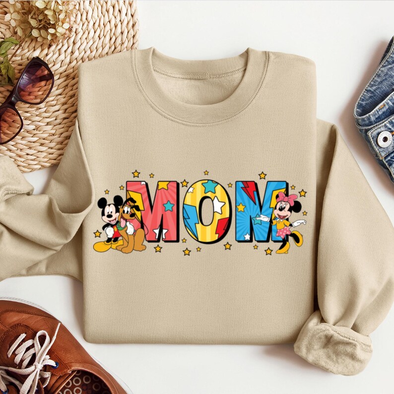 K&ouml;nnte beinhalten: Ein beiger Pullover mit einem grafischen Aufdruck des Wortes "MOM" in bunten Buchstaben. Mickey Mouse und Pluto befinden sich auf der linken Seite des Designs, und Minnie Mouse auf der rechten Seite.