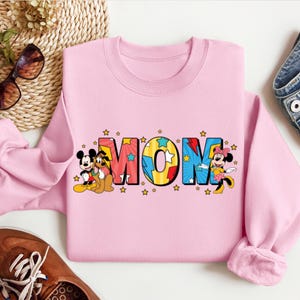 K&ouml;nnte beinhalten: Rosafarbener Pullover mit einem Aufdruck von Mickey Mouse und Pluto, die neben dem Wort "MOM" in bunten Comic-Buch-Lettern mit Sternen stehen.