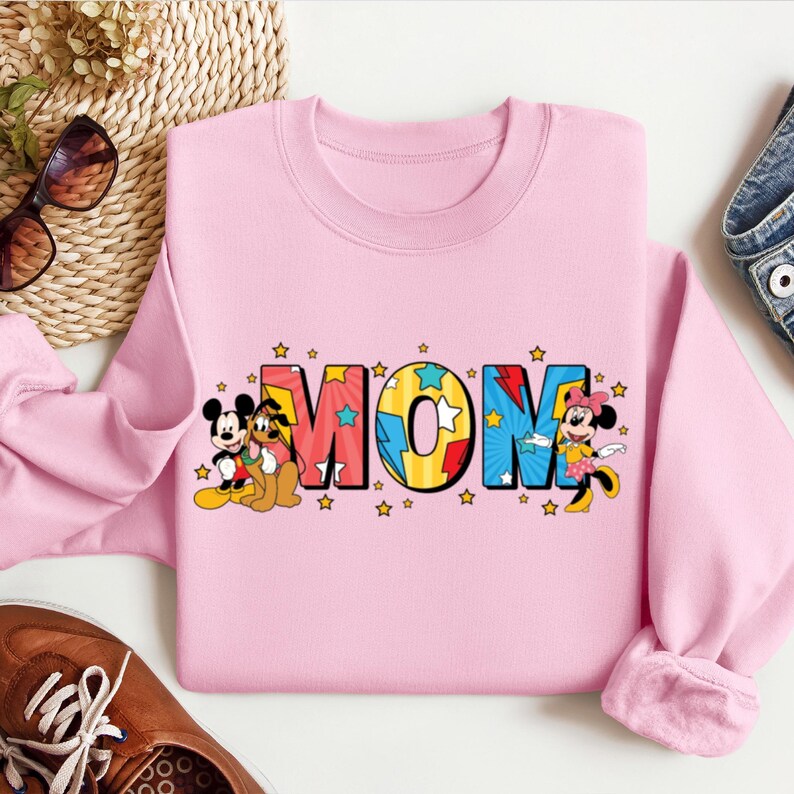 K&ouml;nnte beinhalten: Rosafarbener Pullover mit einem Aufdruck von Mickey Mouse und Pluto, die neben dem Wort "MOM" in bunten Comic-Buch-Lettern mit Sternen stehen.