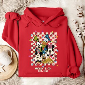 May include: Red hooded sweatshirt with a Christmas-themed graphic featuring Mickey Mouse, Minnie Mouse, Donald Duck, Goofy, and Pluto. The graphic includes a checkered background with string lights and the text "Mickey & Co. Est. 1928".