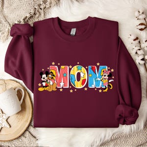 K&ouml;nnte beinhalten: Burgunderfarbener Pullover mit einer bunten Grafik von Mickey Mouse, Minnie Mouse und Pluto, die das Wort "MOM" mit gelben Sternen buchstabieren.