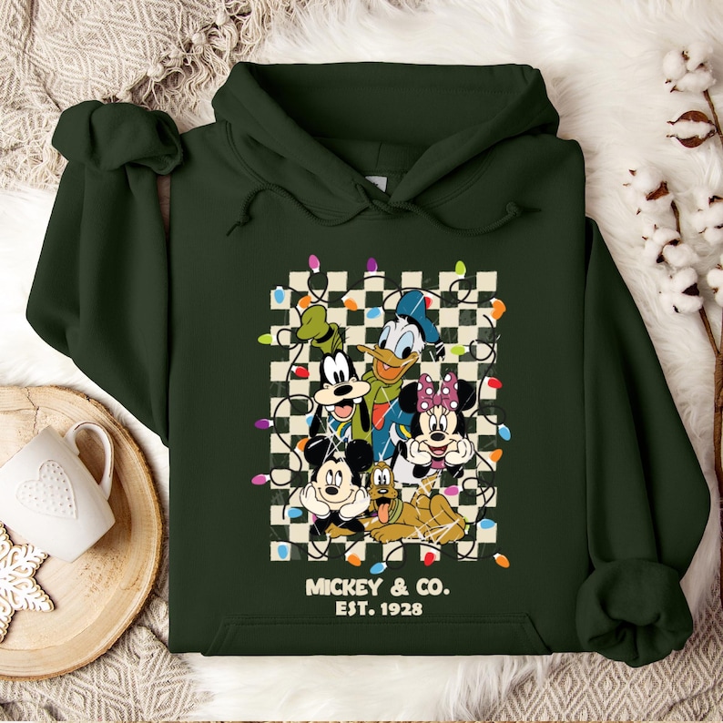 May include: Green hooded sweatshirt with a checkered background featuring Mickey Mouse, Donald Duck, Goofy, Pluto, and Minnie Mouse. The characters are surrounded by colorful Christmas lights. The text "Mickey & Co. Est. 1928" is printed below the image.