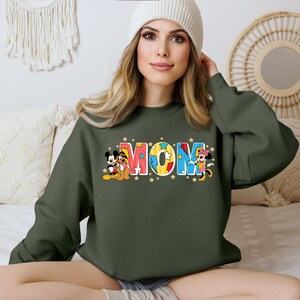 K&ouml;nnte beinhalten: Ein gr&uuml;ner Pullover mit einer bunten "MOM"-Grafik mit Mickey Mouse, Minnie Mouse und Pluto. Die Grafik ist von gelben Sternen umgeben.