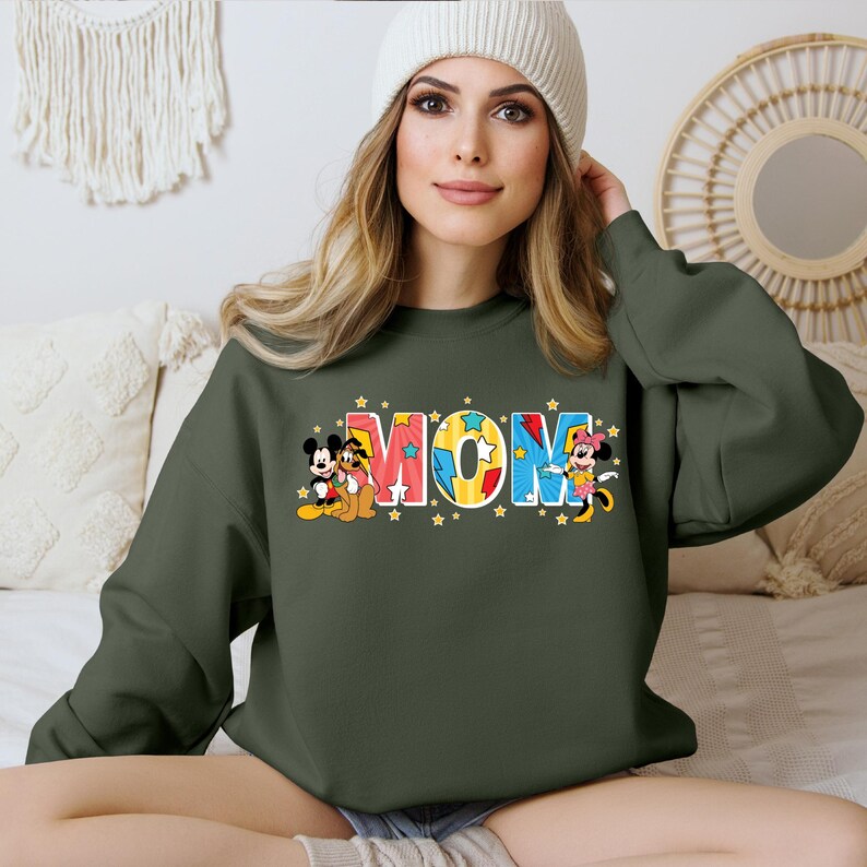 K&ouml;nnte beinhalten: Ein gr&uuml;ner Pullover mit einer bunten "MOM"-Grafik mit Mickey Mouse, Minnie Mouse und Pluto. Die Grafik ist von gelben Sternen umgeben.