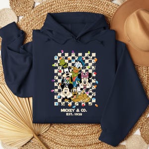 May include: Navy blue hoodie with a checkered background featuring Mickey Mouse, Donald Duck, Goofy, Pluto, and Minnie Mouse. The characters are surrounded by colorful Christmas lights. The text "Mickey & Co. Est. 1928" is printed below the characters.