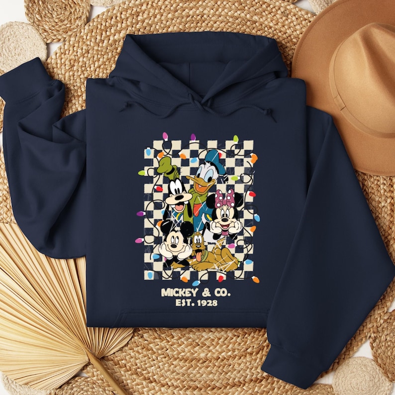 May include: Navy blue hoodie with a checkered background featuring Mickey Mouse, Donald Duck, Goofy, Pluto, and Minnie Mouse. The characters are surrounded by colorful Christmas lights. The text "Mickey & Co. Est. 1928" is printed below the characters.
