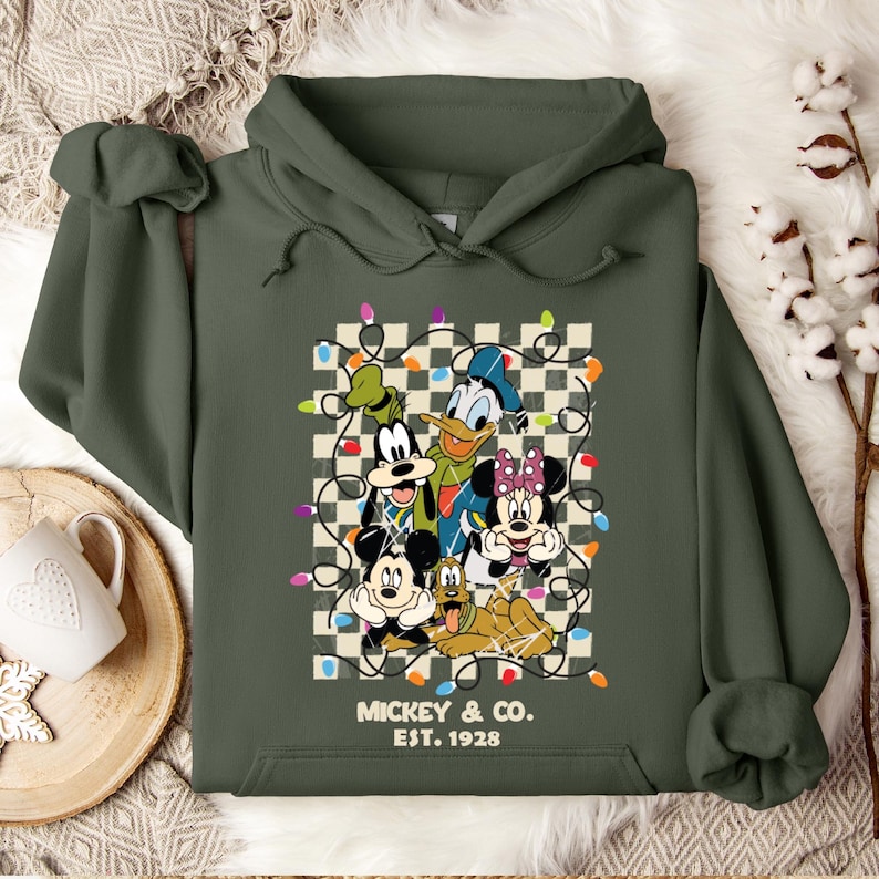 May include: Green hooded sweatshirt with a vintage-style graphic of Mickey Mouse, Donald Duck, Goofy, Minnie Mouse, and Pluto. The graphic features the characters in front of a checkered background with colorful Christmas lights. The text "Mickey & Co. Est. 1928" is below the graphic.