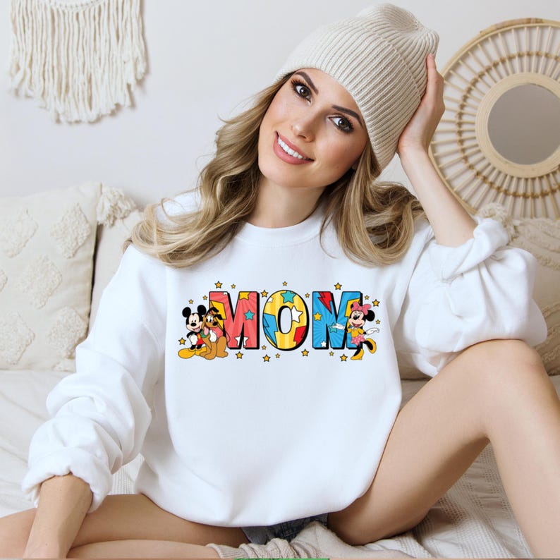 K&ouml;nnte beinhalten: Wei&szlig;er Pullover mit einem bunten "MOM"-Motiv mit Mickey Mouse, Minnie Mouse und Pluto. Die Buchstaben sind mit gelben Sternen umrandet.